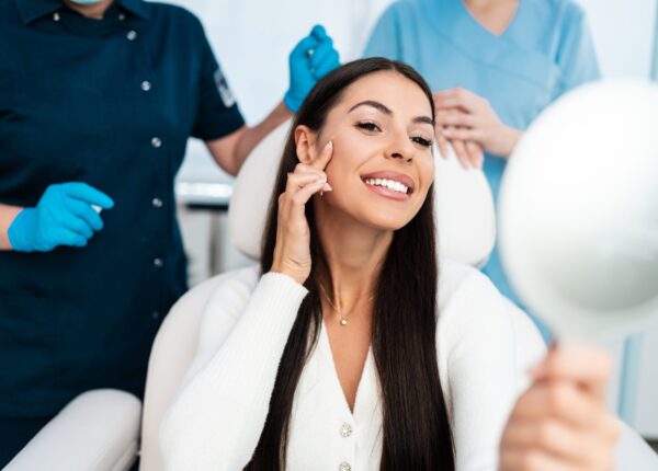 Beautiful and happy young woman sitting in medical chair and looking in the mirror. She is satisfied after successful beauty treatment with hyaluronic acid fillers or botulinum toxin injections.