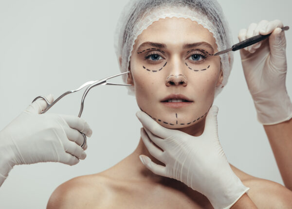 Close up of woman having cosmetic face surgery with scalpel and medical forceps against grey background. Woman having a anti aging and face lift surgery.