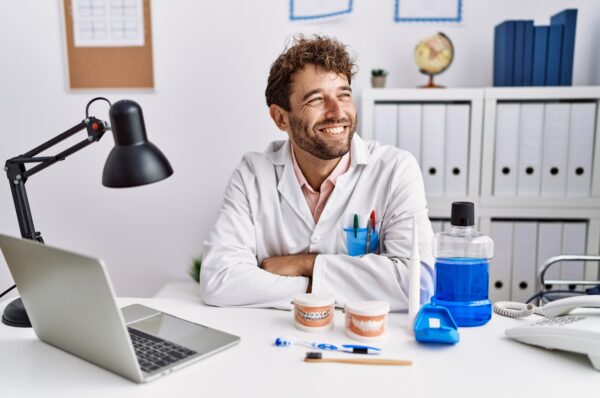 Young hispanic dentist man working at medical clinic looking away to side with smile on face, natural expression. laughing confident.