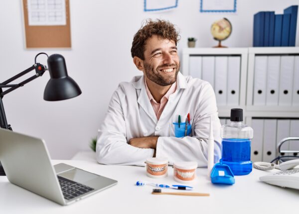 Young hispanic dentist man working at medical clinic looking away to side with smile on face, natural expression. laughing confident.